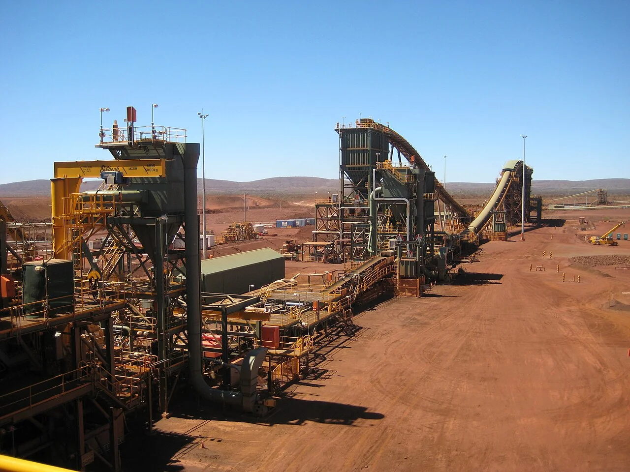 Fotografía referencial: torres de procesamiento de mineral, tolvas, pasarelas y correas transportadoras en una planta minera de paisaje árido.