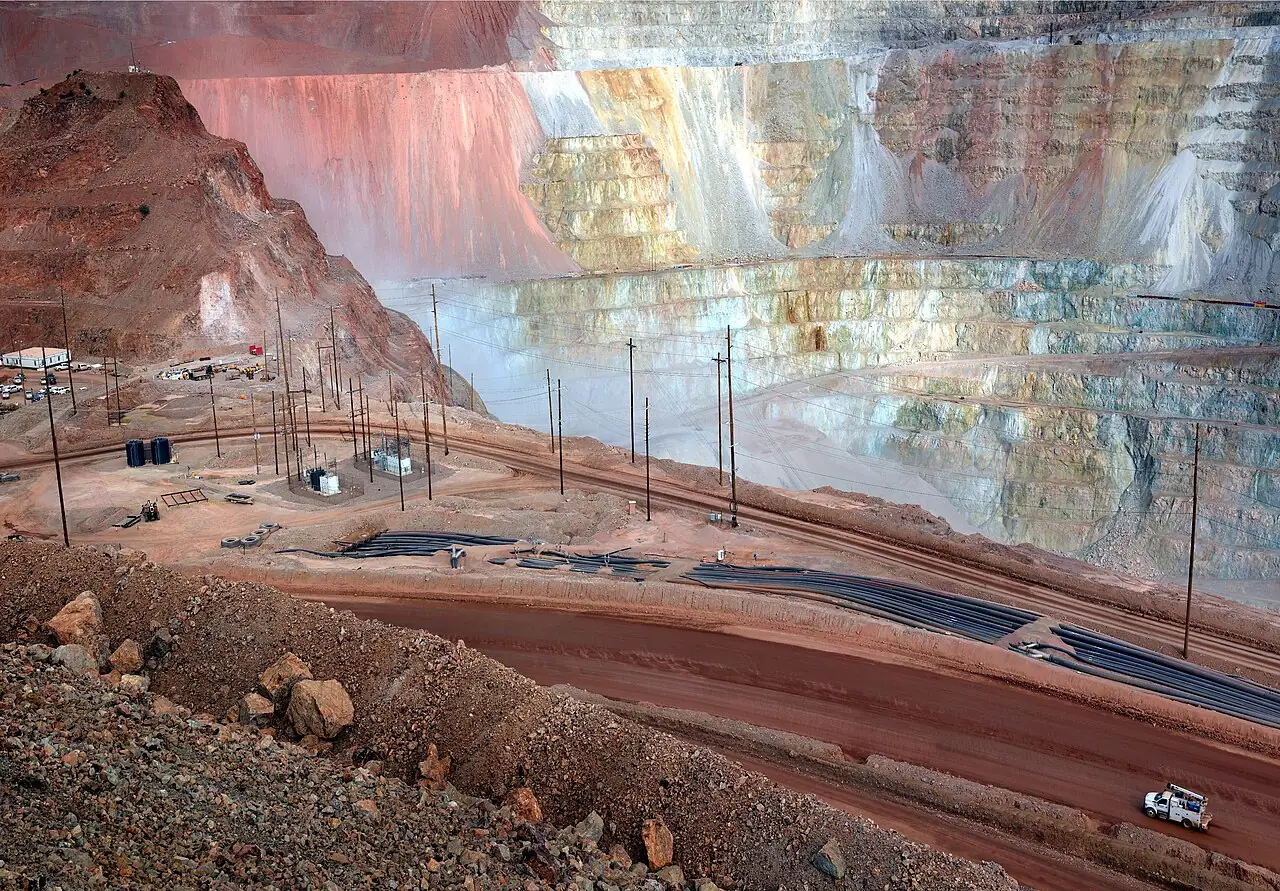 Fotografía referencial: caminos de operación, tuberías, instalaciones eléctricas y vehículos de apoyo junto a los bancos de una mina de cobre a cielo abierto en terreno desértico.